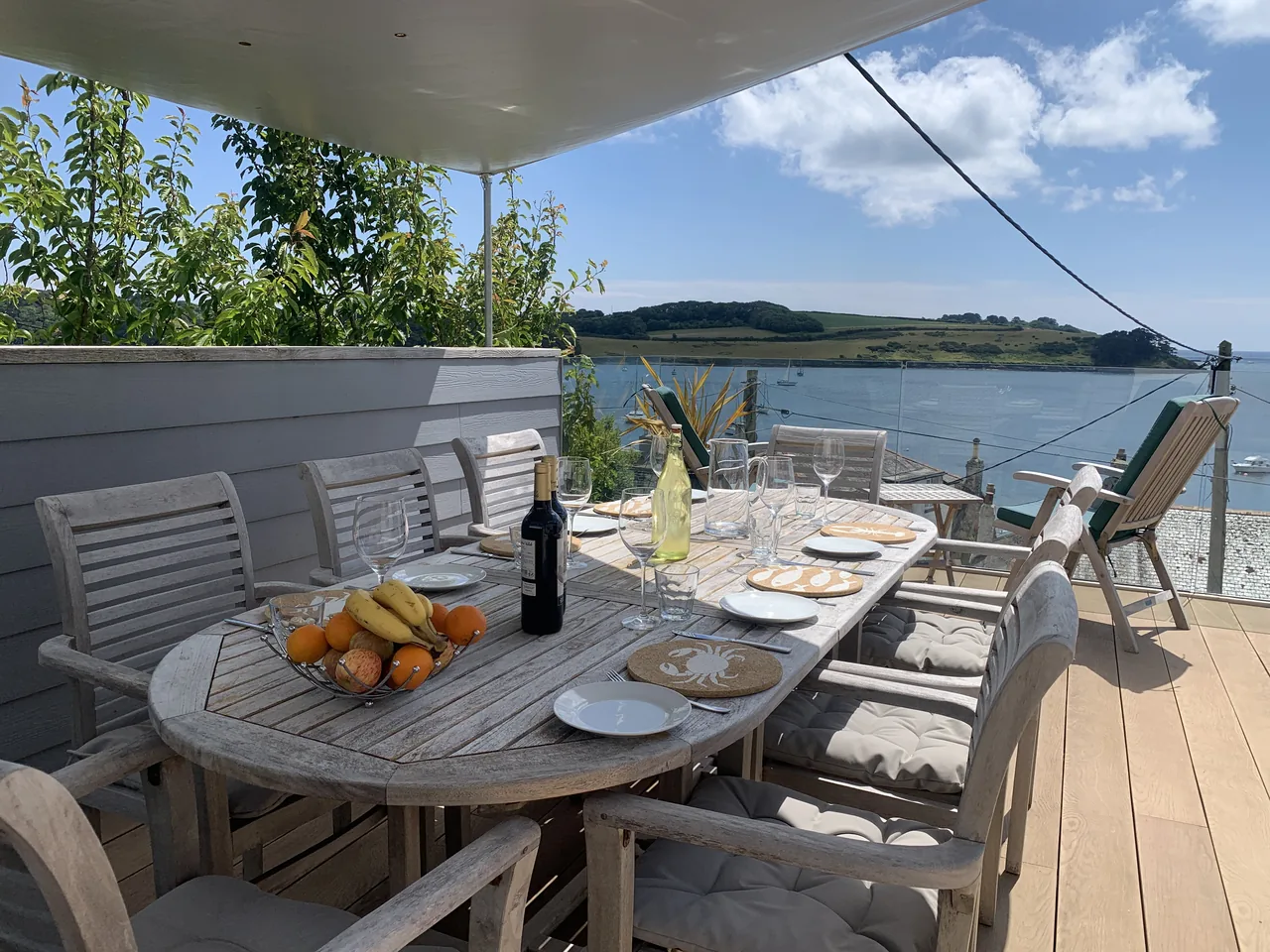 A table set on the wooden terrace beneath a shade sail, with the water and green headland beyond.